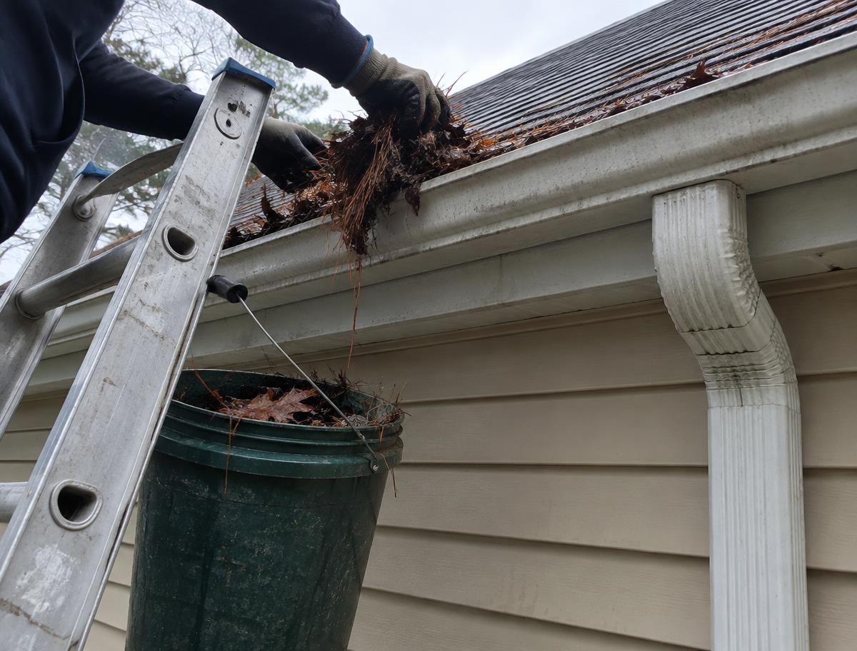 Gloved hands clearing soaked leaves out of a rain gutter from a ladder
